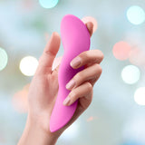 A hand with neatly manicured nails holds a pink, curved, and smooth object against a blurred background with colorful bokeh lights. The ergonomic design of the FemmeFunn Unda Panty Vibrator Pink ensures it fits comfortably in your hand.