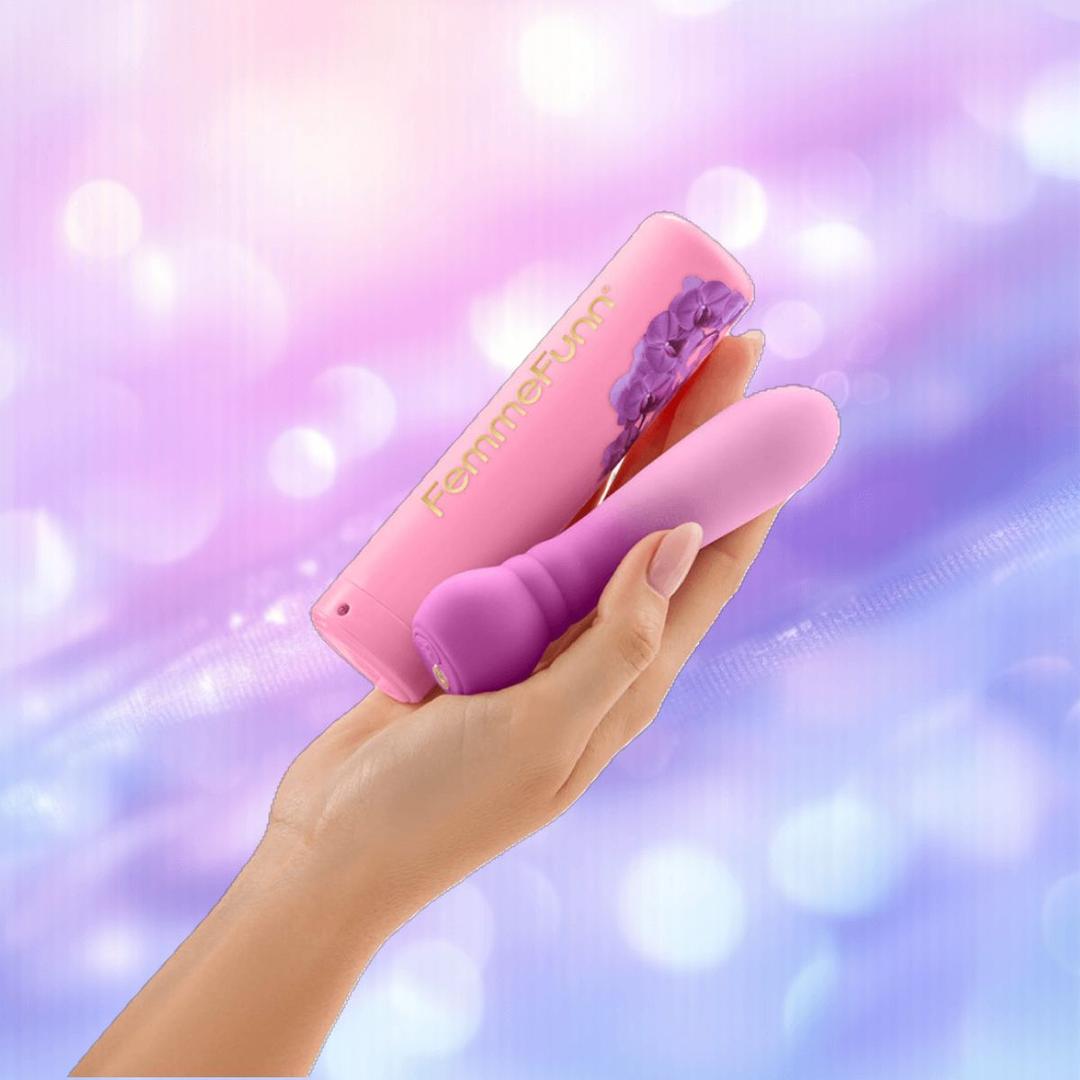 A hand holds the FemmeFunn Anniversary Ultra Bullet Orchid vibrator and its matching Femme Funn case against a soft, purple-blue bokeh background.