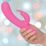 A hand holds the CalExotics First Time Rechargeable Bunny with Turbo Boost Accelerated Speeds, set against a softly blurred pastel background, hinting at powerful dual stimulation and intense pleasure.