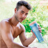 A shirtless young man with short brown hair sits outdoors, holding a Fleshlight Fleshjack Boys Allen King: Realistic Male Masturbator Majestic Cake Butt in his right hand and looking at the camera, with greenery blurred in the background.