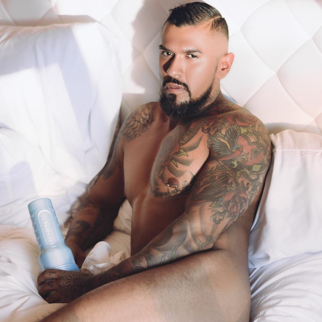 A muscular, tattooed man with a beard sits nude on a bed with white sheets, holding the Fleshlight Fleshjack Boys Boomer Banks: Realistic Male Masturbator Cake Butt, looking directly at the camera.