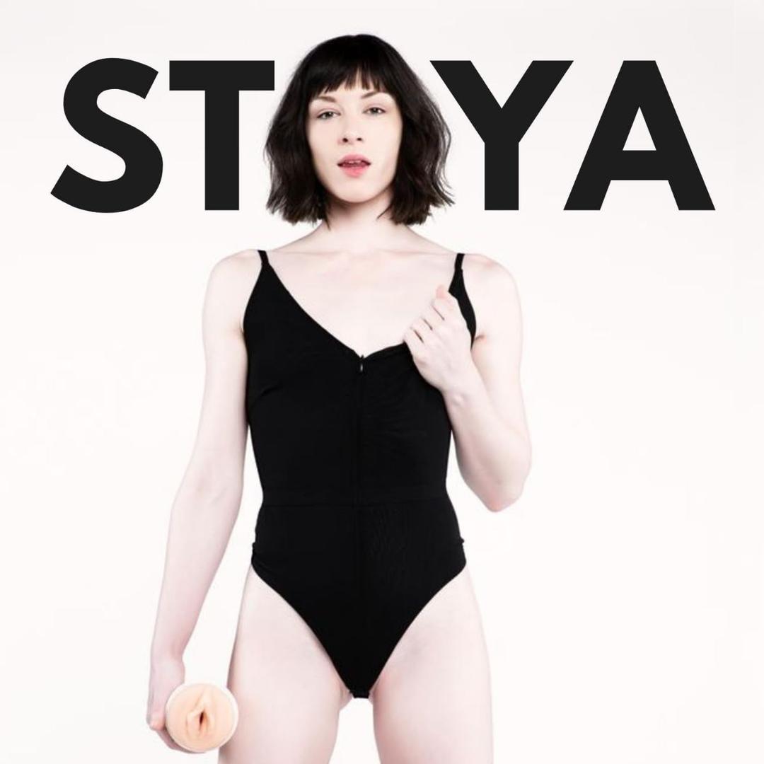 A pale-skinned, dark-haired person in a black bodysuit stands holding an object, with "STOYA" in bold behind them, inspired by Fleshlight Girls: Stoya Destroya, available at Condomania.com.