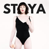 A pale-skinned, dark-haired person in a black bodysuit stands holding an object, with "STOYA" in bold behind them, inspired by Fleshlight Girls: Stoya Destroya, available at Condomania.com.