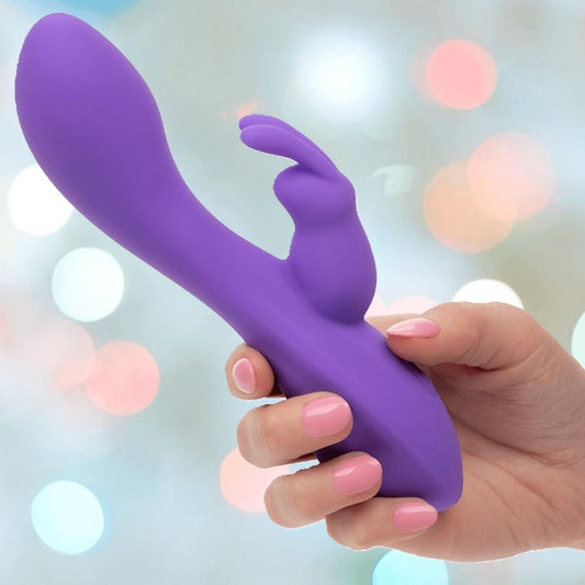A hand with pink-painted nails holds the CalExotics Flex Bend & Hold Double Rumbler Rabbit Vibrator—featuring a curved shaft and bunny-shaped stimulator—against a soft, blurred background. 1080