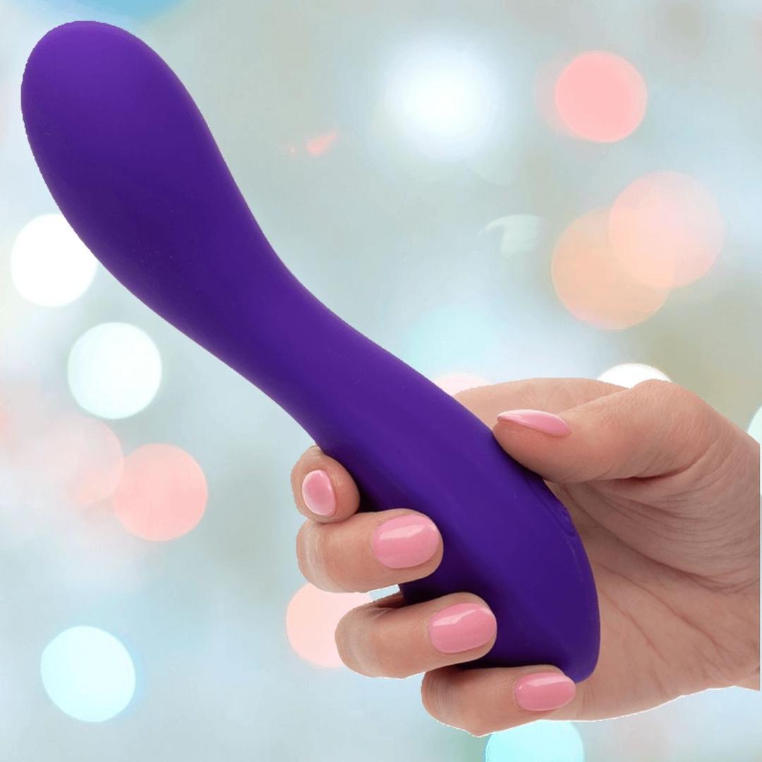 A hand with light pink nail polish holds the purple Flex Bend & Hold Rumbler G-Spot Stimulator by CalExotics, set against a backdrop of soft, colorful bokeh lights.