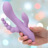 A hand with pink nails holds the CalExotics Flex Bend & Hold Triple Rumbler, a purple silicone vibrator with multiple attachments, against a softly blurred pastel background.