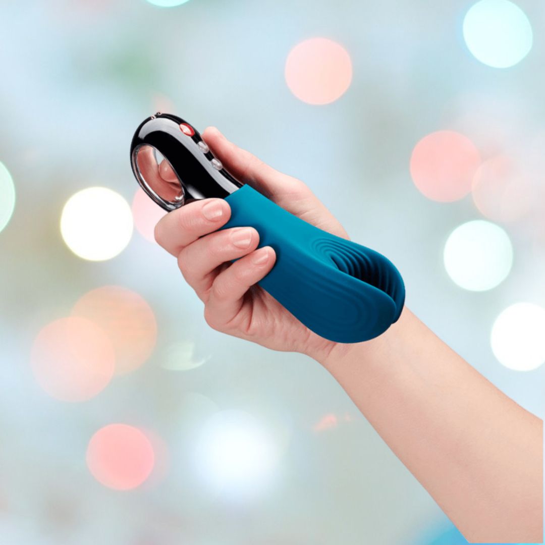 A person is holding the Fun Factory Manta Rechargeable Vibrating Male Stroker for Men in Deep Blue against a vibrant, blurred background. The device features a sleek design with convenient controls on one end, perfect for a personalized experience.