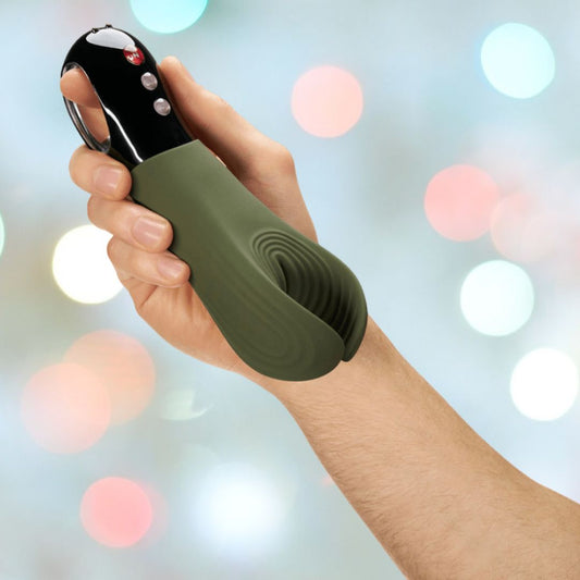 A hand holding the Fun Factory Manta Rechargeable Vibrating Male Stroker For Men in Moss Green, a silicone gadget with a metallic handle and buttons, set against a backdrop of colorful, blurred lights. 1080