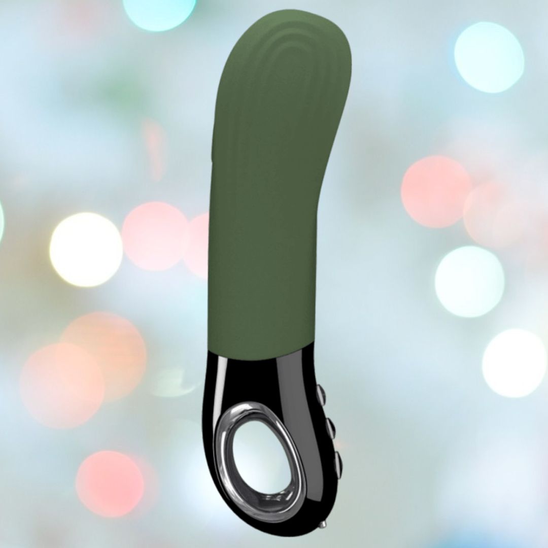 The Fun Factory Manta Rechargeable Vibrating Male Stroker for Men in Moss Green stands against a blurred background with pastel bokeh lights. Featuring a curved design and three-button controls on the handle, this versatile personal massager is perfect for both solo and couple play, offering an ideal blend of style and function.