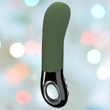 The Fun Factory Manta Rechargeable Vibrating Male Stroker for Men in Moss Green stands against a blurred background with pastel bokeh lights. Featuring a curved design and three-button controls on the handle, this versatile personal massager is perfect for both solo and couple play, offering an ideal blend of style and function.