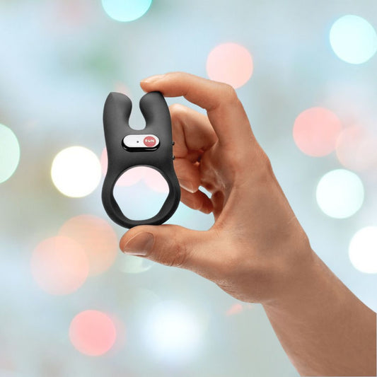 A hand holds the Fun Factory NOS Vibrating Silicone Cock Ring - Black, showcasing its U-shaped silicone design against a blurred background with colorful bokeh lights, and promising hands-free orgasms with its dual-motor vibration feature. 1080