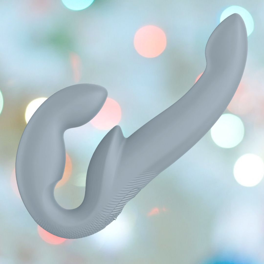 The Fun Factory Share Vibe Pro Cool - Grey, a gray, dual-ended couples vibrator with a curved design, is displayed against a blurry background with pastel-colored circular lights and promises strong vibrations for mutual pleasure.