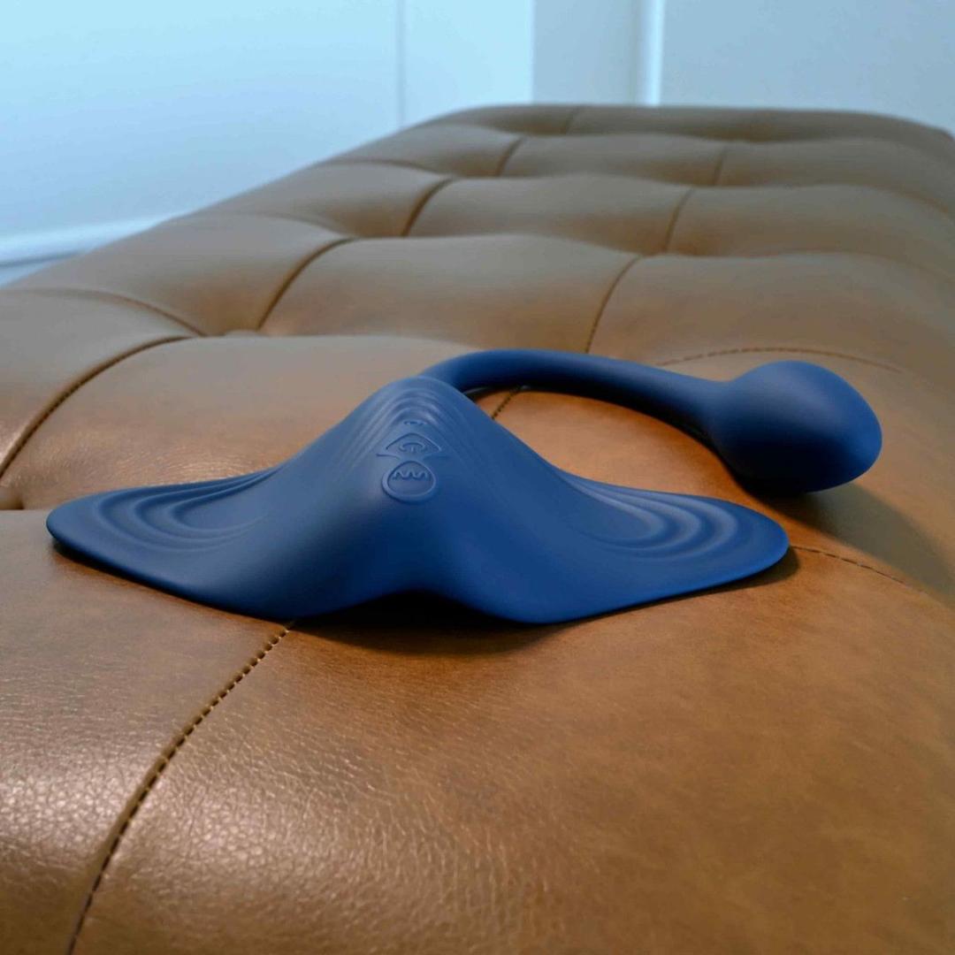 A blue silicone Gender X Daily Grind vibrating plug by Evolved Novelties, with a long bulbous end and curved edges, rests on a tan leather cushion. The body-safe toy features subtle embossed patterns on its flat base.