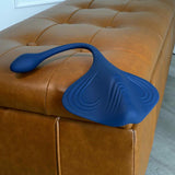 The Gender X Daily Grind by Evolved Novelties, a blue silicone vibrating plug with a flexible stem and textured surface, rests on a tan tufted leather ottoman. Its curved lines and buttons emphasize its modern, body-safe design.