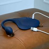 A navy blue silicone curved vibrating plug, Gender X Daily Grind by Evolved Novelties, with a small bulb at one end, is charging via USB cable on a brown leather ottoman.