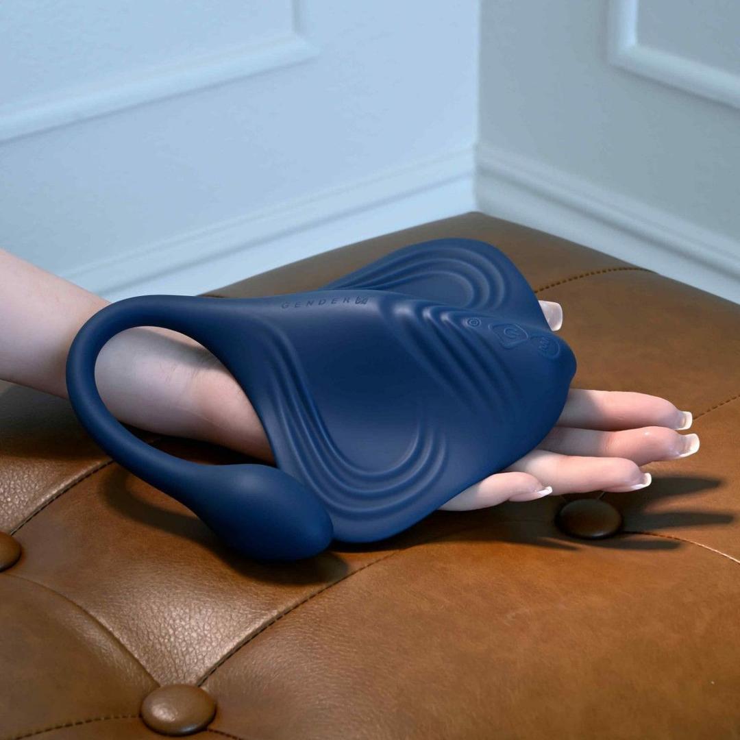 A hand holds the Gender X Daily Grind by Evolved Novelties, a blue silicone, body-safe vibrating plug with textured ridges, resting on a brown leather cushion in a room with white paneled walls.