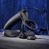 The Evolved Novelties Gender X Full Swing, a black silicone dual motor vibrator ring and prostate massager, charges via USB on a wooden surface with black stones against a blue wooden backdrop.