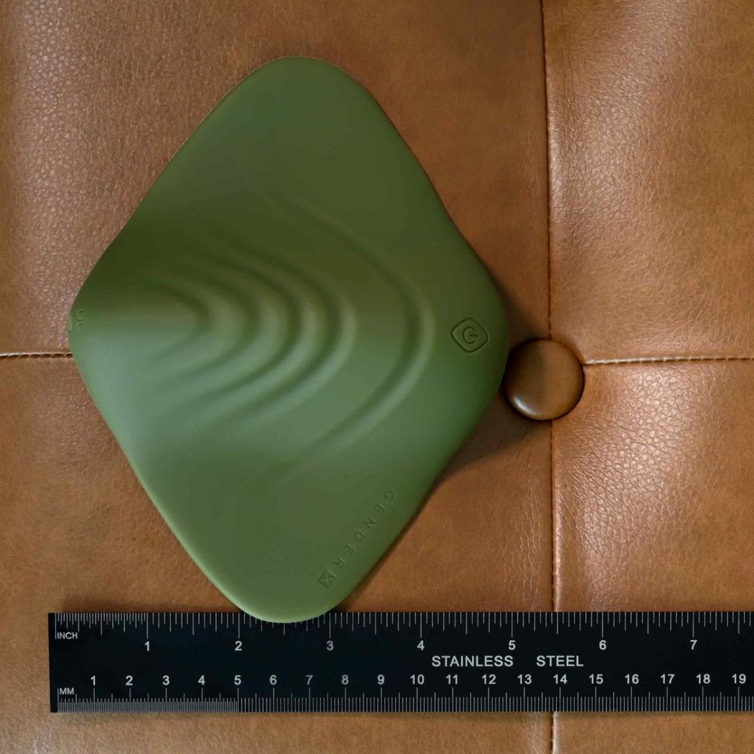 The Evolved Novelties Gender X Pleasure Pad is a green, textured, square vibrating toy with rounded edges, wave patterns, and a "G" button. Shown about 6 inches wide on tan leather beside a ruler, it's designed for all genders.
