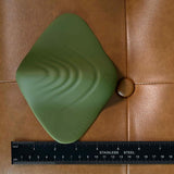 The Evolved Novelties Gender X Pleasure Pad is a green, textured, square vibrating toy with rounded edges, wave patterns, and a "G" button. Shown about 6 inches wide on tan leather beside a ruler, it's designed for all genders.