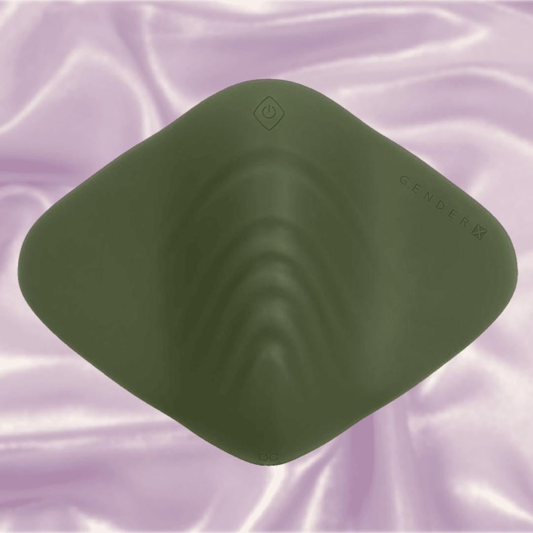 The Gender X Pleasure Pad by Evolved Novelties is a dark green, diamond-shaped silicone pad shown on soft pink satin for a gender-inclusive vibe.
