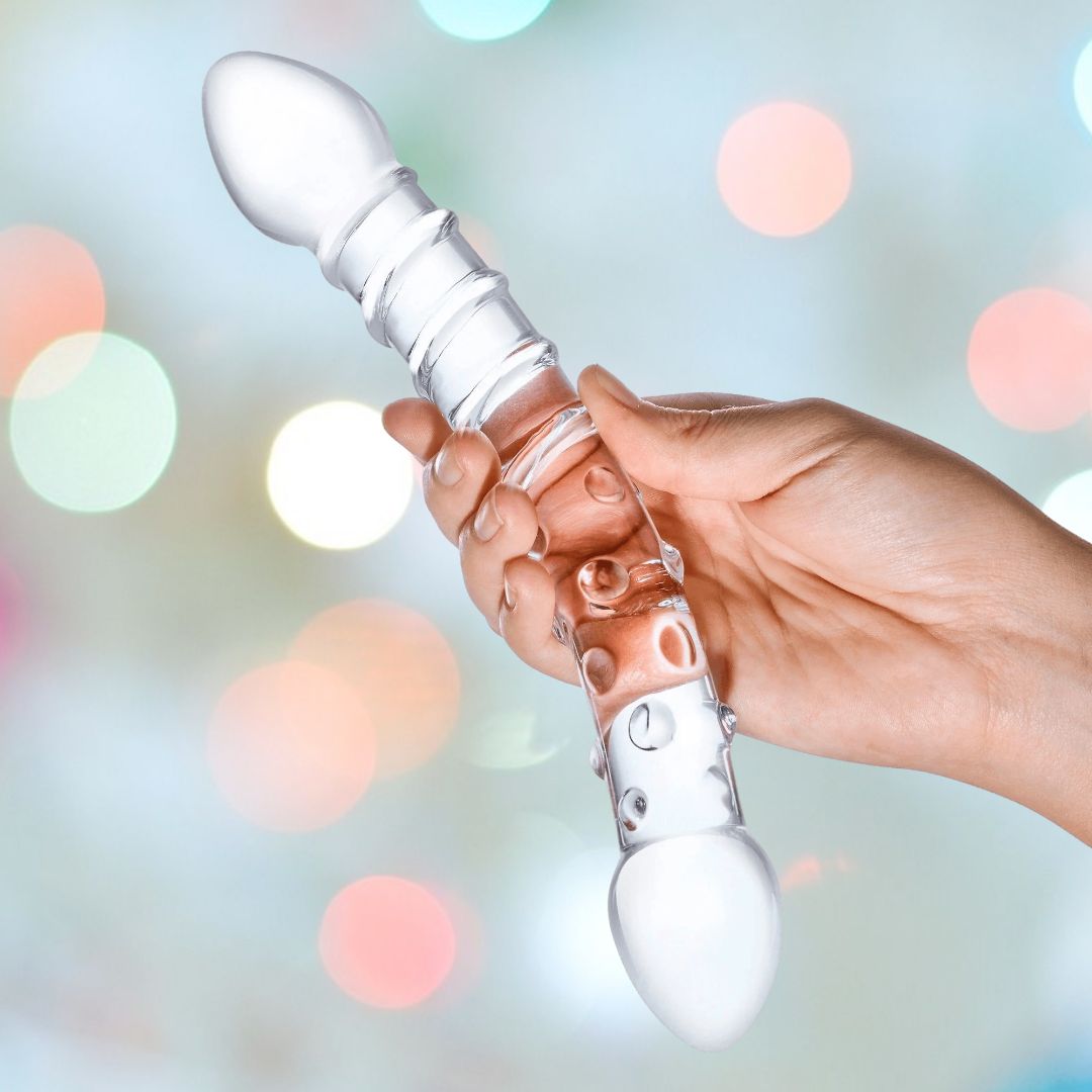 A hand holds the Gläs 10" "Double Trouble" Double-Sided Glass Dildo, which boasts a clear design and textured handle, complete with dual-ended spherical ends ideal for G-spot or P-spot massage. In the background, vibrant, blurred lights create a festive ambiance.