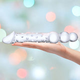 A hand holds the Gläs 12" Double Ended Glass Dildo, featuring a transparent and textured design with a smooth, rounded end for versatile use. The background showcases soft pastel-colored bokeh lights.