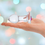 A hand holds the Gläs 3.5" "Bling Bling" Glass Butt Plug, showcasing its hypoallergenic design with a bulbous end and flat base, set against a softly blurred background adorned with colorful bokeh lights.