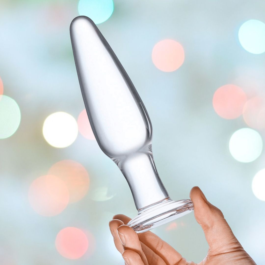 A hand holding a transparent object from the Gläs 3 Piece Anal Training Set against a softly blurred backdrop featuring multicolored lights. The piece from the Gläs set has a tapered design with a rounded base.