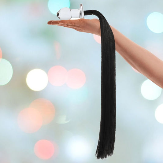 A hand delicately holds the Gläs 3" Horse Tail Glass Butt Plug, featuring a sleek, long black tassel, against a softly blurred background where bokeh lights shimmer in pastel colors. 1080