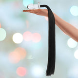 A hand delicately holds the Gläs 3" Horse Tail Glass Butt Plug, featuring a sleek, long black tassel, against a softly blurred background where bokeh lights shimmer in pastel colors.