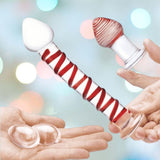 Hands hold items from the Gläs Mr. Swirly Set: a clear glass reminiscent of a dildo, a smooth white-tipped piece, and a red-striped object on a stand. Two small rounded glass Kegel balls rest in an open palm against a blurred backdrop with lights.