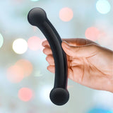 A hand holds the Gläs 5.5" Double Bull Black Glass Dildo, a sleek, hypoallergenic object with round ends, against a softly blurred background of colorful lights.