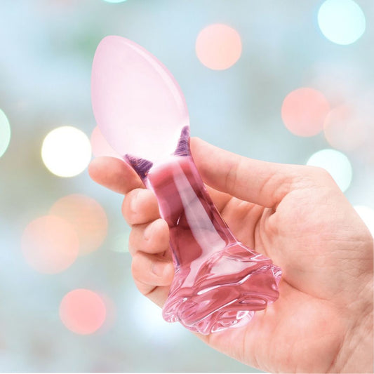 A hand cradles a translucent, pink glass piece—similar to the Gläs 5" Rosebud Glass Butt Plug—featuring a rounded, spoon-like tip and an intricate, flower-inspired base. The soft blur of pastel hues and light circles in the background sets an ideal ambiance for temperature play exploration. 1080