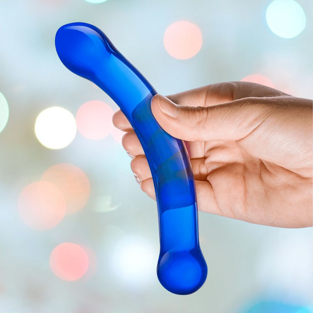 A hand elegantly grips the Gläs 6" Curved G-Spot Blue Glass Dildo, masterfully designed for optimal G-Spot stimulation. The softly blurred background shimmers with multi-colored bokeh lights, enhancing the allure of this captivating product from Gläs.