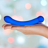 A hand holding the Gläs 6" Curved G-Spot Blue Glass Dildo, crafted with elegantly rounded ends for optimal G-spot stimulation. The softly blurred background features bokeh lights in a range of pastel colors.