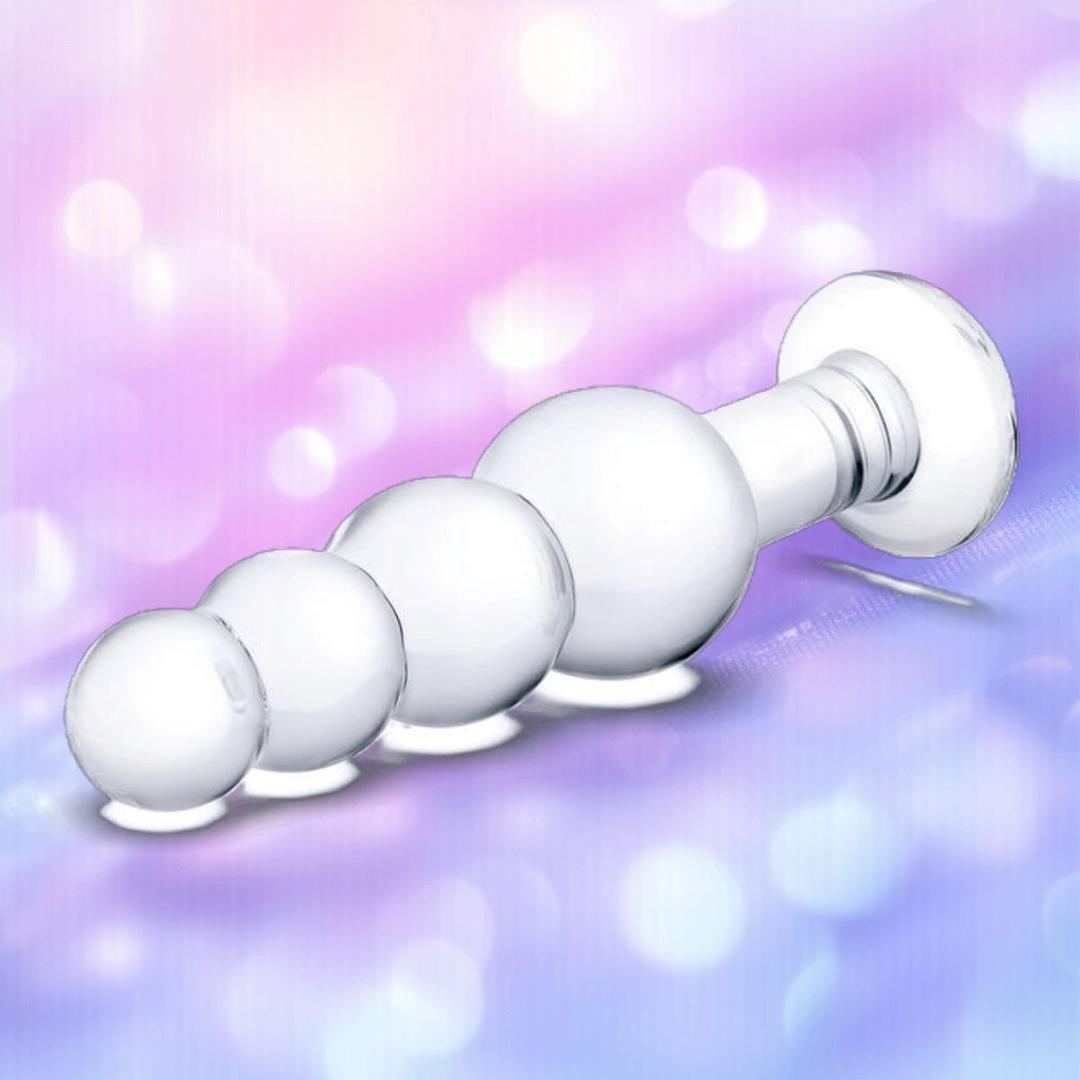 The Gläs 7.25" Glass Beaded Butt Plug by Gläs features four smooth, body-safe glass beads in ascending size—ideal for temperature play and displayed against a soft, pastel background with gentle bokeh lights.