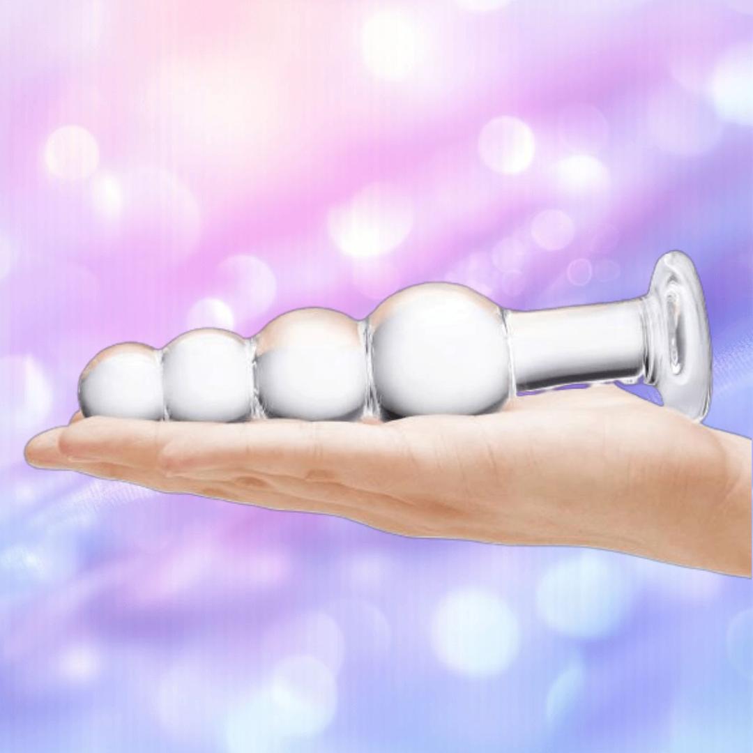 A hand holds the Gläs 7.25" Glass Beaded Butt Plug by Gläs, with its flared base visible against a soft pastel background in pink and purple hues with bokeh light effects.