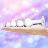 A hand holds the Gläs 7.25" Glass Beaded Butt Plug by Gläs, with its flared base visible against a soft pastel background in pink and purple hues with bokeh light effects.