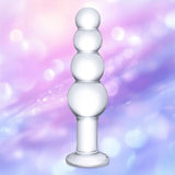 The Gläs 7.25" Glass Beaded Butt Plug by Gläs features four stacked glass beads and a flat base, shown upright against a pastel pink and purple blurred background—body-safe and perfect for temperature play.
