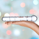 A hand gently holds the Gläs 7" Callisto Clear Glass Dildo, with its smooth, rounded end ready for G-spot exploration. The textured shaft shines against a softly blurred background featuring pastel bokeh in shades of pink, blue, and green.