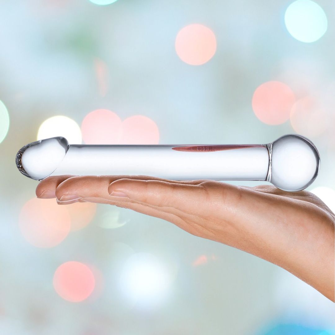 A hand holding the Gläs 7" Realistic Head Glass Dildo, designed with rounded ends ideal for temperature play, is set against a soft-focus background featuring colorful, circular light spots.