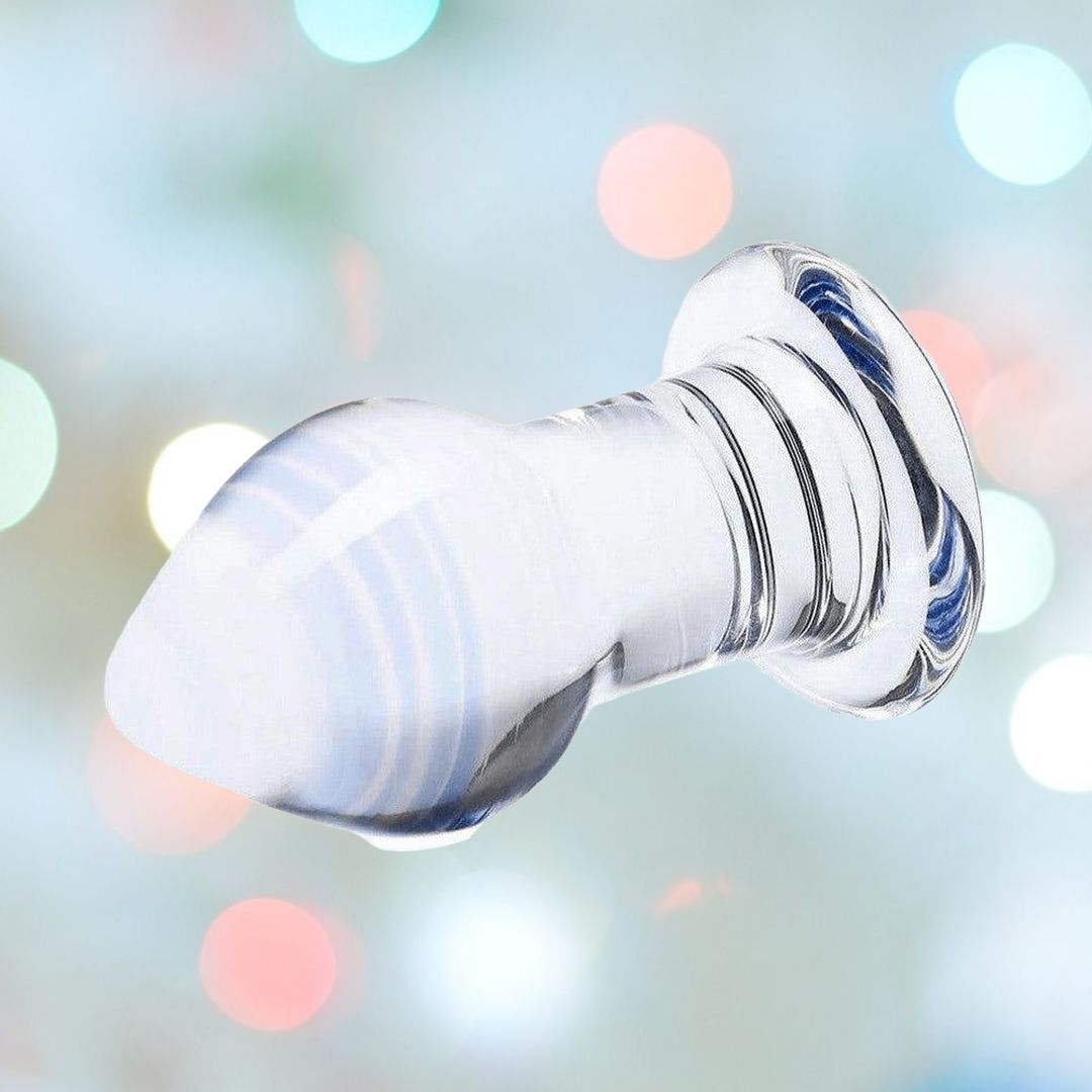 A translucent Gläs Amethyst Rain Glass Butt Plug with a bulbous end and a narrow stem, set against a blurred background of various soft-colored bokeh lights. The plug features a smooth, reflective surface adorned with faint blue streaks.