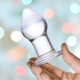 A hand holding the Gläs Amethyst Rain Glass Butt Plug with a flared base. The background showcases a softly blurred array of pastel-colored lights, creating a festive and dreamy atmosphere, perfect for enhancing moments of anal play.