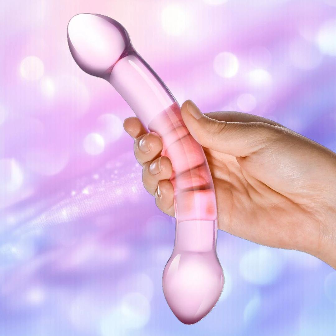 A hand holds the Gläs Double Trouble Purple Dildo by Gläs with curved double ends against a soft, pastel bokeh background—ideal for temperature play and versatile G-spot stimulation.