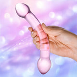 A hand holds the Gläs Double Trouble Purple Dildo by Gläs with curved double ends against a soft, pastel bokeh background—ideal for temperature play and versatile G-spot stimulation.