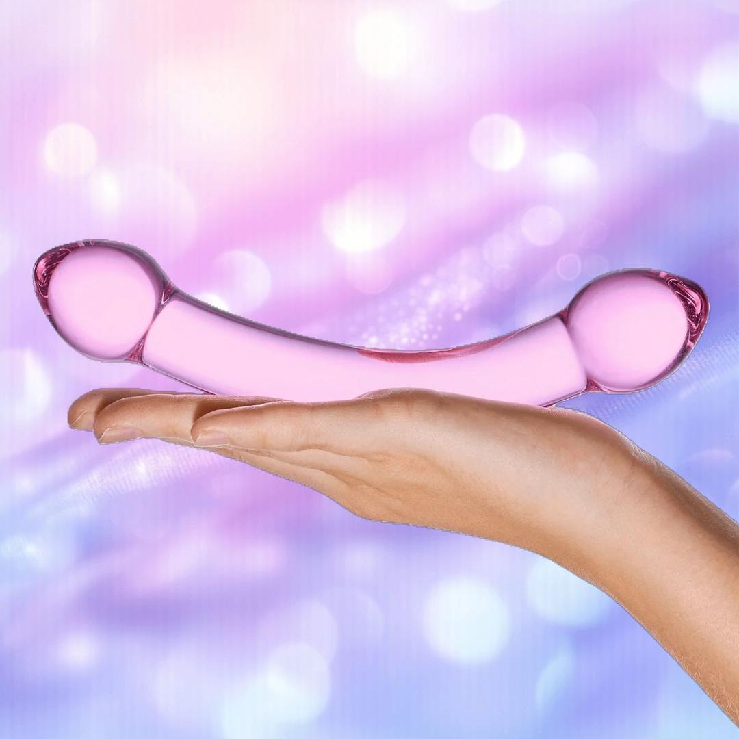 A hand displays the Gläs Double Trouble Purple Dildo by Gläs against a soft, pastel backdrop with blue and purple bokeh effects. This translucent, double-ended glass toy is ideal for temperature play and versatile pleasure.