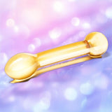 The Gläs Honey Dripper 7" Double Ended Anal Dildo by Gläs, featuring rounded yellow glass ends, lies on a soft-focus pink and purple background.
