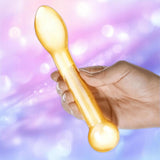 A hand holds the Gläs Honey Dripper 7" Double Ended Anal Dildo by Gläs, a translucent yellow toy with rounded and flat spoon-like ends, set against a softly blurred purple and blue background.