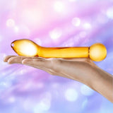 A hand holds the Gläs Honey Dripper 7" Double Ended Anal Dildo—a smooth, translucent yellow glass toy by Gläs with rounded ends—set against a soft pastel pink and purple bokeh background.