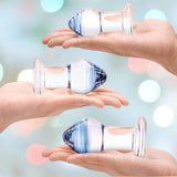 The Glas Pleasure Droplets Anal Training Kit from Gläs, featuring three glass items with blue accents, is showcased on open hands against a pastel backdrop with pink and green bokeh effects.